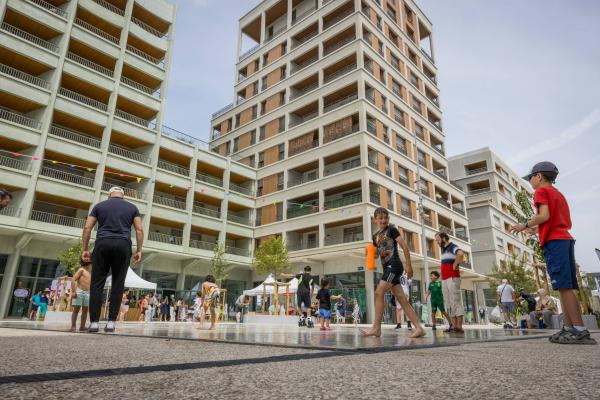 Children and adults play and walk near an urban splash pad in a city plaza, surrounded by modern mid-rise apartment buildings.
